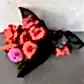 A vibrant bouquet of red roses and pink and orange dahlias from Flowers on Norton St, the Strawberry Kisses Mixed Bouquet 2024, is wrapped in black paper beside a small bag of red "Strawberry Kisses" candy against a gray textured wall and surface.