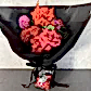 The "Strawberry Kisses Mixed Bouquet" by Flowers on Norton St provides a whimsical display with pink and red roses, purple and orange flowers in black paper. A small bag of red candies, called Strawberry Kisses, is tied with a black ribbon, set against a plain, light-colored wall.