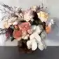 The Peaches + Cream Mixed Flower Vase by Flowers on Norton St features Colombian roses, Cloud Moth orchids, peach and cream roses, pink dahlias, white orchids, and dark wispy foliage in a textured brown vase. It beautifully contrasts a light gray background.