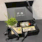 A black gift box from the "Flowers on Norton St" brand, part of the Royal Botanical Gardens Fragrance Collection, includes a small potted plant reminiscent of those at the gardens, a bottle, hand cream, and two decorative bars. The box is lined with black crinkle paper and adorned with a black ribbon featuring the words "FLOWERS ON NORTON.