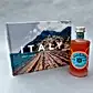 The "An Amalfi Moment Gift Box" by Flowers on Norton St features a book titled "ITALY" by Gray Malin, highlighting La Dolce Vita with vibrant beach scenes, alongside a bottle of Italian-made Malfy Gin Rosa known for its refreshing grapefruit flavor, all set against a simple gray surface.