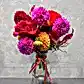 Introducing "Lovely Vintage Vases" by FlowersonNortonSt—a vibrant arrangement in a clear glass vase, ideal for a table centerpiece. This exquisite bouquet showcases pink dahlias, red roses, orange blossoms, and an array of other colorful seasonal flowers. It's elegantly tied with a pink and orange ribbon, set against a plain light gray wall.