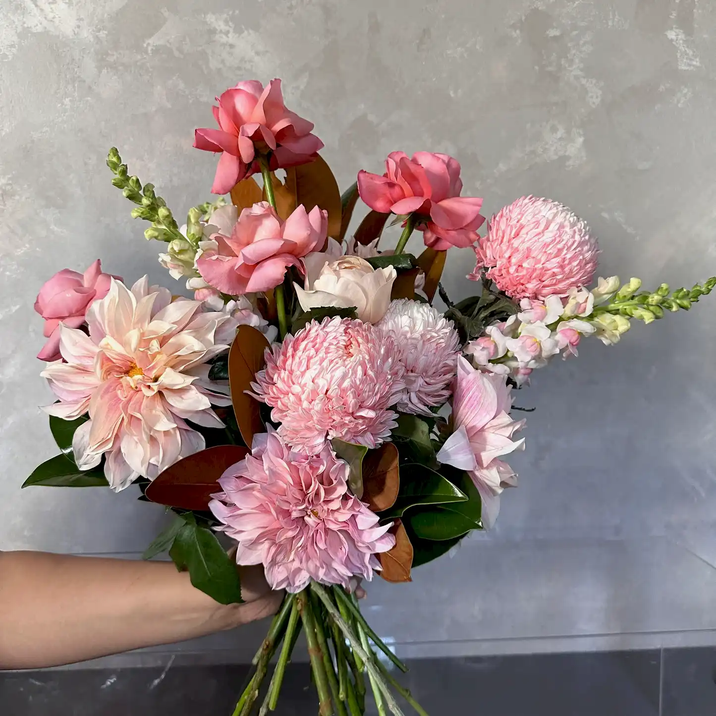 A hand holds the Pink Expression Seasonal Bouquet 2024 by Flowers on Norton St, featuring pink roses, dahlias, and seasonal blooms with lush green leaves, set against a textured gray background.