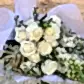 The Cream Classic Mixed Flowers by FlowersonNortonSt features a beautiful arrangement of white roses, dahlias, and elegant greenery with delicate white and green fillers. The white and cream blooms are wrapped in pristine white paper, creating a stunning display on the wooden surface.