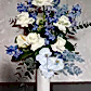 The "Botanical Blues Mixed Flower Vase" by FlowersonNortonSt features an artistic arrangement of white roses, blue orchids, and anthuriums in a tall white vase. This unique bouquet includes a mix of blue and white flowers complemented by lush greenery, all beautifully set against a light gray textured background.