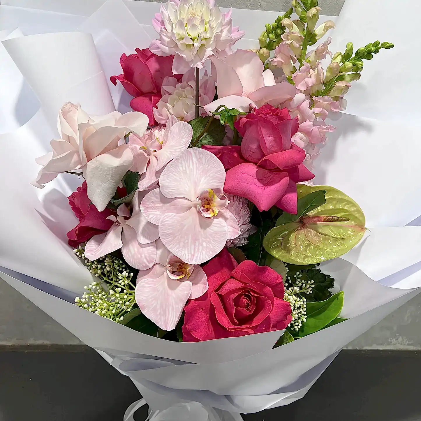 Introducing "Tickle me Pink" by FlowersonNortonSt: A stunning handcrafted botanical bouquet featuring a mix of pink and white flowers such as orchids and roses, alongside other unique blooms, all beautifully wrapped in white paper. This arrangement showcases an array of pink and green hues, offering a vibrant and sophisticated display that makes for an exceptional gift on any occasion.