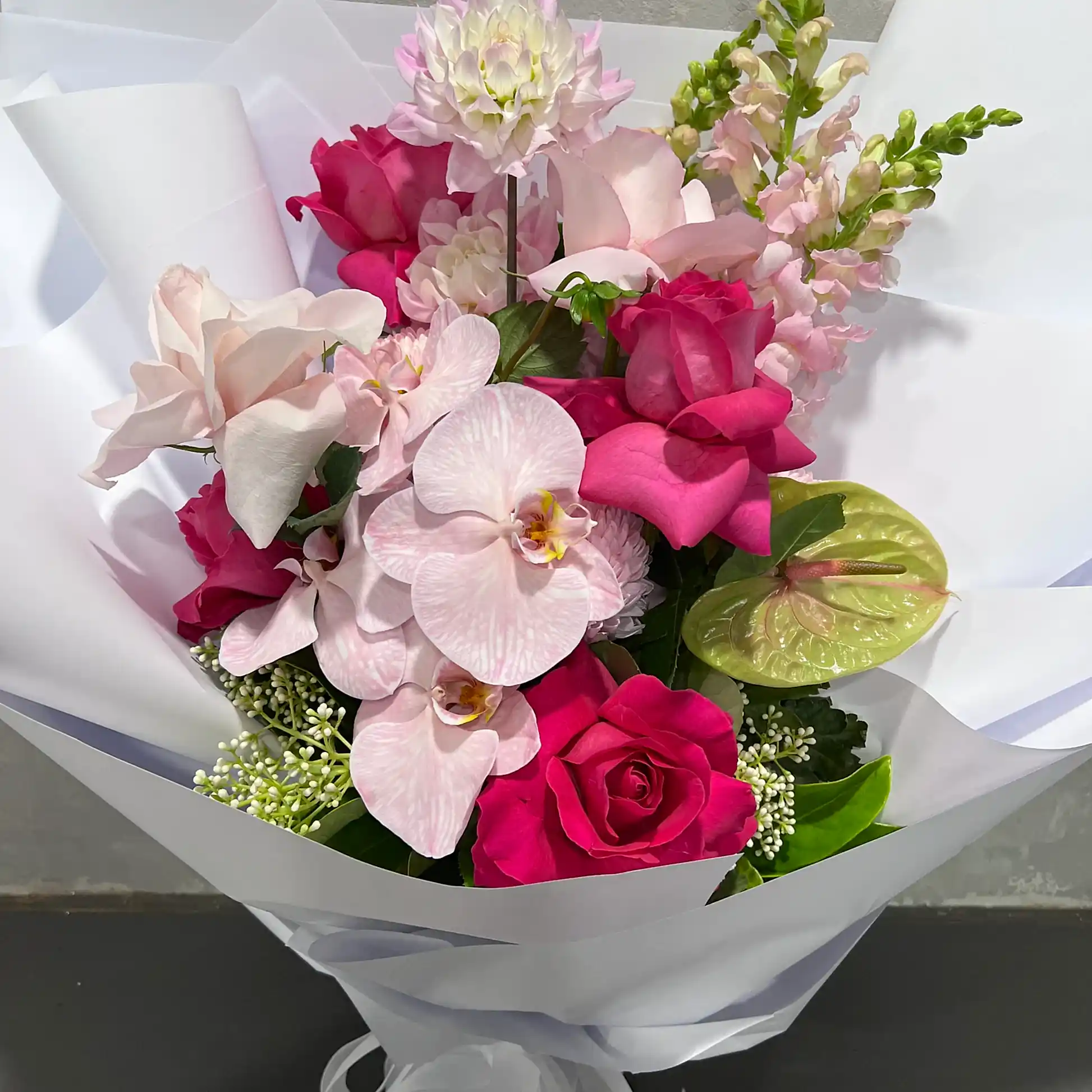 Introducing "Tickle me Pink" by FlowersonNortonSt: A stunning handcrafted botanical bouquet featuring a mix of pink and white flowers such as orchids and roses, alongside other unique blooms, all beautifully wrapped in white paper. This arrangement showcases an array of pink and green hues, offering a vibrant and sophisticated display that makes for an exceptional gift on any occasion.