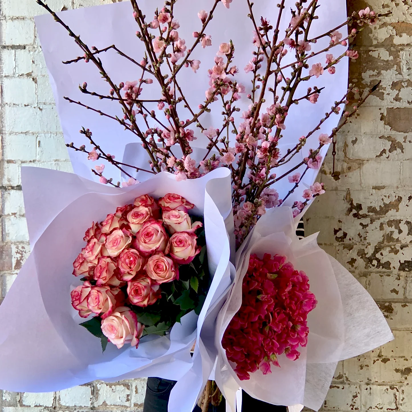 The "Colour Me Pink Seasonal Flowers" arrangement by Flowers on Norton St features peach blossoms at the center, flanked by a bouquet of seasonal pink and white roses on one side and bright pink hydrangeas on the other, all elegantly wrapped in white paper set against a textured brick wall.
