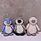 Three Penguin Plush Toys by FlowersonNortonSt, in pastel shades of blue, gray, and pink, are standing side by side against a textured wall backdrop. Each toy features a similar design with dark beak accents and tiny feet.