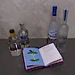 Four bottles from the Vodka Gift Set by Flowers on Norton St are arranged on a table next to an open cocktail recipe book, showcasing an "Apple Martini" recipe with an illustration of an apple. The background displays a plain concrete wall.