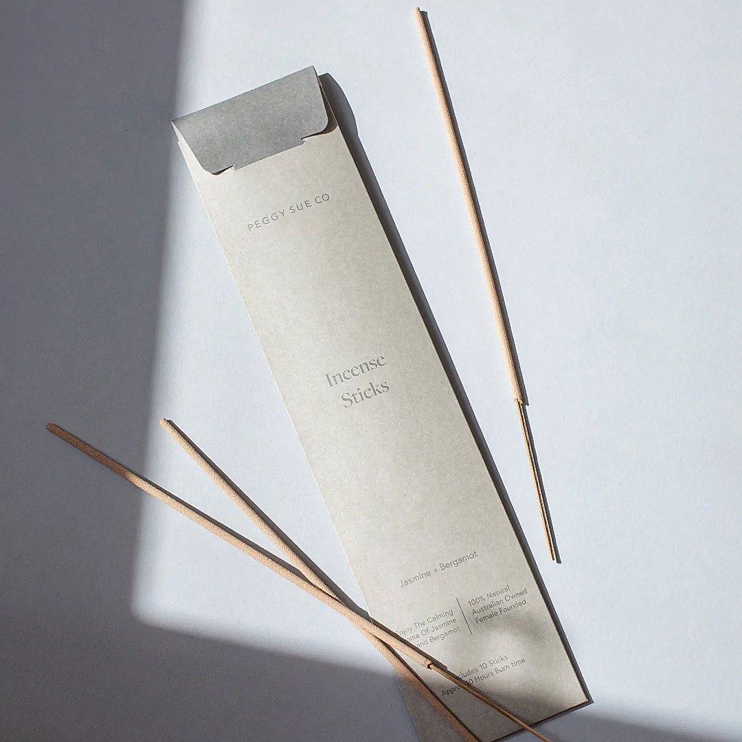 A package labeled "Incense Sticks | Peggy Sue Co." by Flowers on Norton St rests on a light surface. Four sandalwood incense sticks are scattered nearby, while sunlight casts a shadow across part of the scene, creating a serene and minimalist composition.