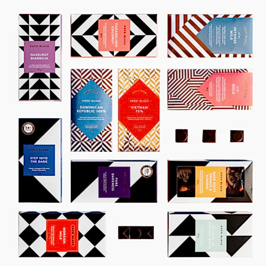 The Koko Black Chocolate Block by FlowersonNortonSt is presented in packaging adorned with geometric patterns and vibrant colors, arranged neatly in a grid. These handcrafted chocolates are made with care, using sustainably sourced cocoa. Some boxes are open, offering a glimpse of the delicious pieces within.