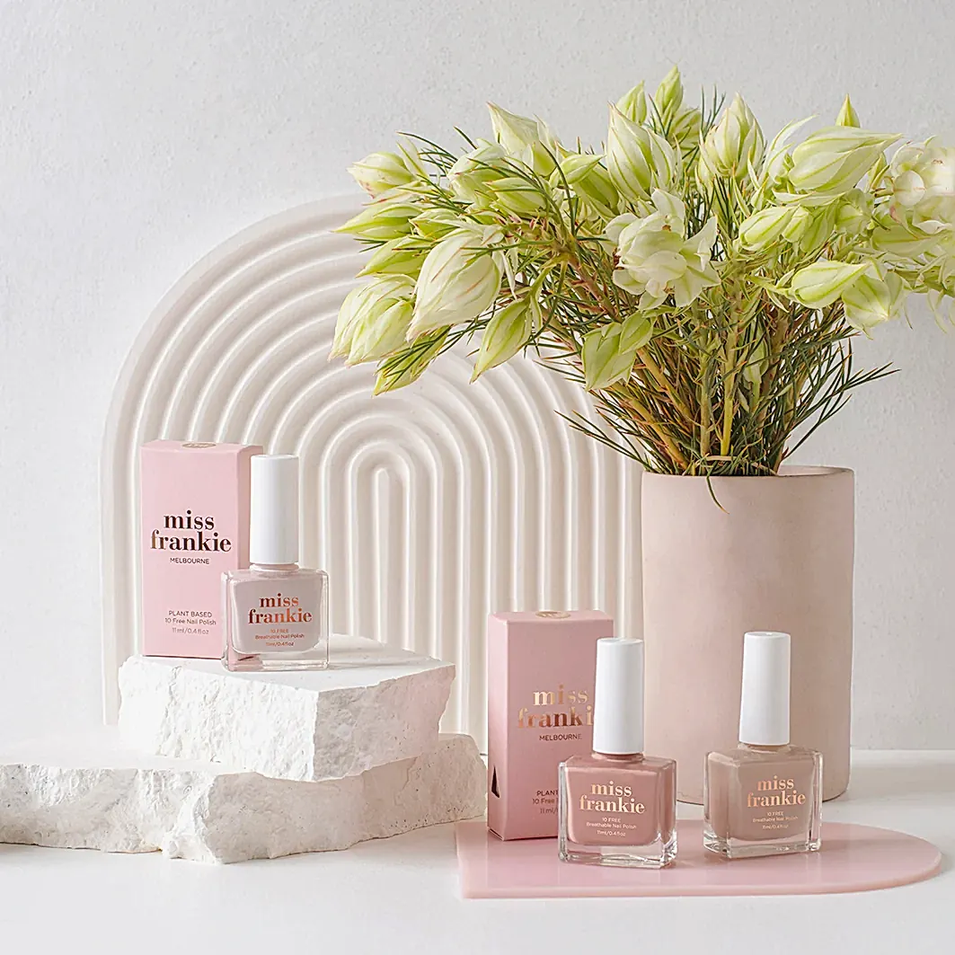 A chic arrangement showcases a pair of MISS FRANKIE | Nail Polish bottles from Flowers on Norton St, elegantly placed on textured white stones and complemented by pink packaging. Adjacent to them is a minimalist vase filled with white flowers, all set against an arched white backdrop.