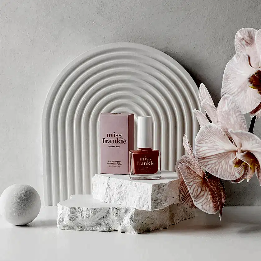 A bottle of MISS FRANKIE Nail Polish by Flowers on Norton St, along with its box, is artfully displayed on textured white stones. The composition features a white arched backdrop and pink orchids that add elegance to the scene. The light pink, chip-resistant nail polish harmoniously matches the hue of the orchids and the packaging.