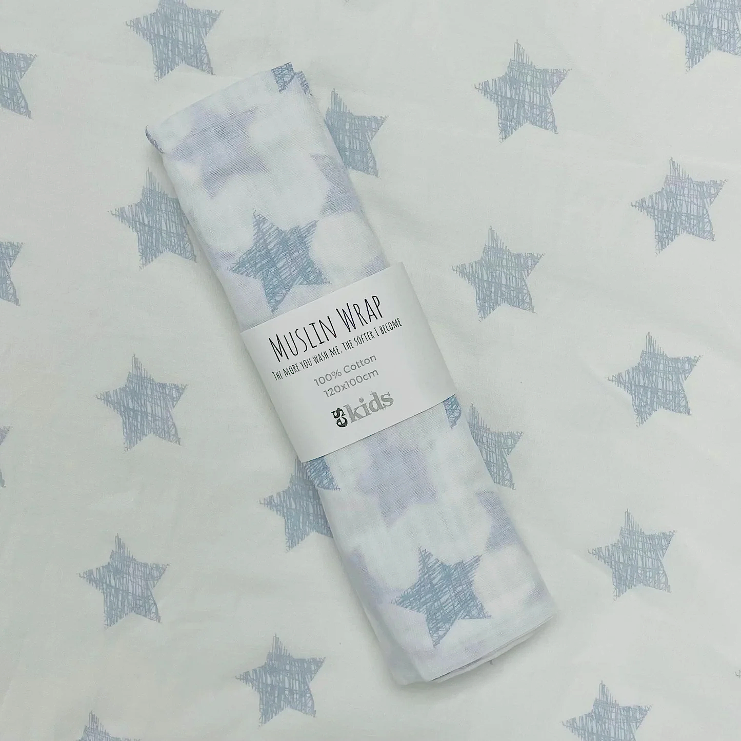 A rolled muslin wrap from FlowersonNortonSt is elegantly placed on a fabric background adorned with star patterns, enhancing the celestial theme. This soft muslin, featuring a matching star design, includes a label that reads "Muslin Wraps" and "100% cotton," making it ideal for baby wrapping.