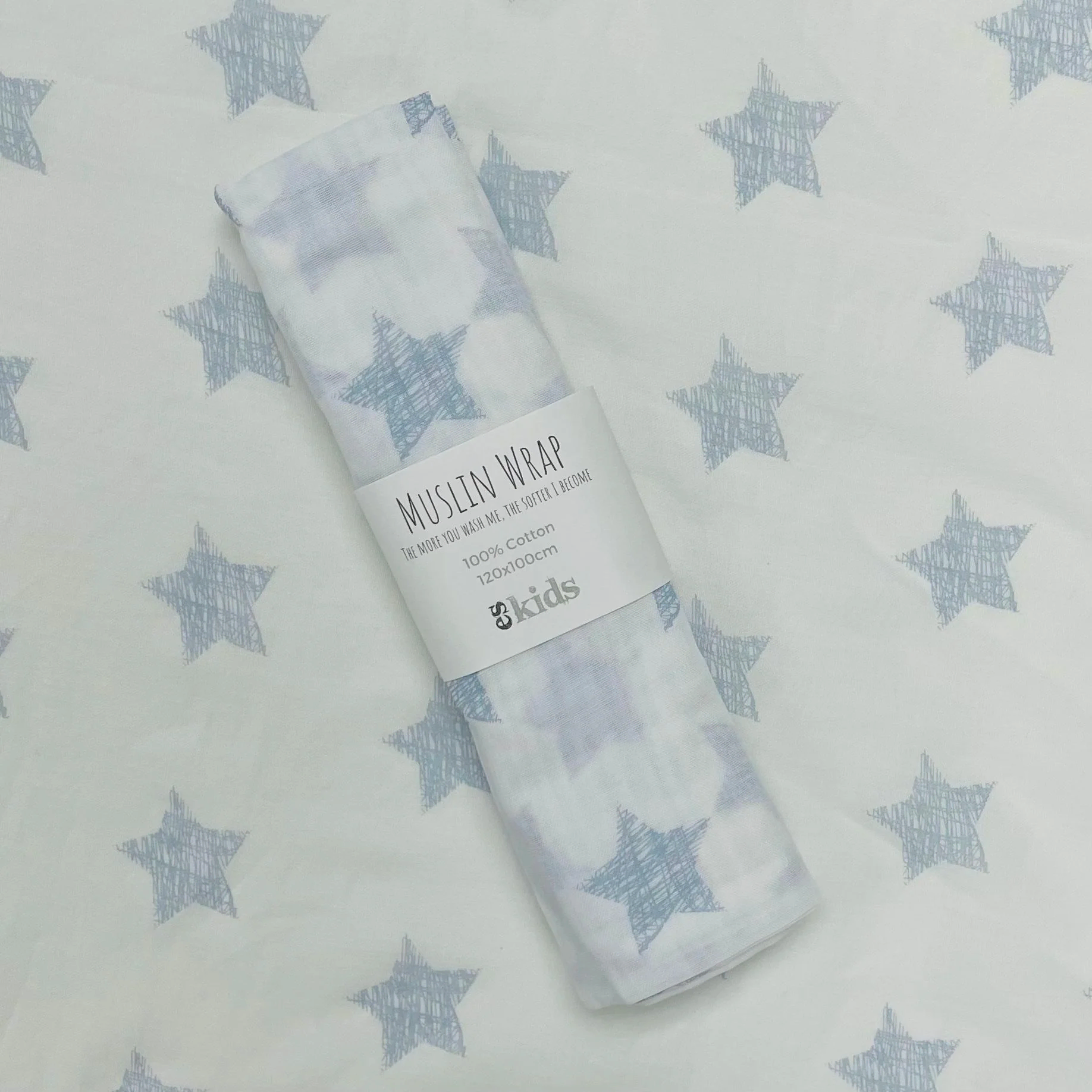 A rolled muslin wrap from FlowersonNortonSt is elegantly placed on a fabric background adorned with star patterns, enhancing the celestial theme. This soft muslin, featuring a matching star design, includes a label that reads "Muslin Wraps" and "100% cotton," making it ideal for baby wrapping.