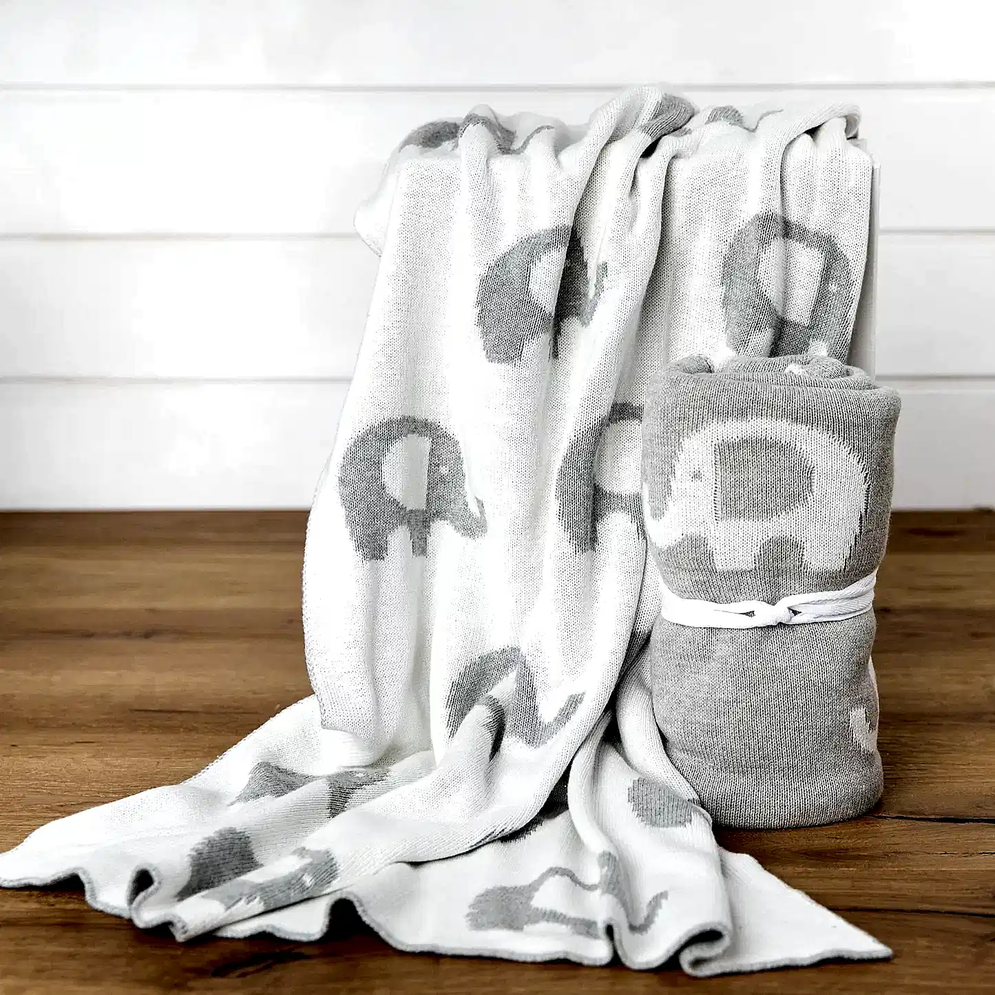 On a wooden surface, two cozy Ellie Baby Blankets from FlowersonNortonSt with adorable gray and white elephant patterns are displayed. One blanket is draped over a ledge, while the other is rolled up and tied with a white ribbon, highlighting their soft texture.