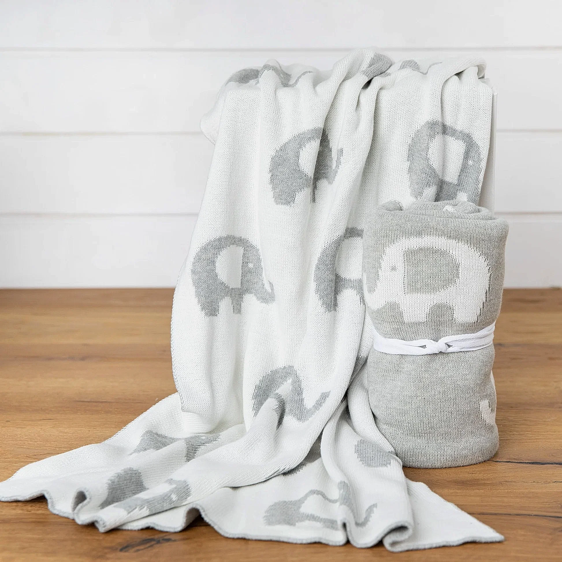 On a wooden surface, two cozy Ellie Baby Blankets from FlowersonNortonSt with adorable gray and white elephant patterns are displayed. One blanket is draped over a ledge, while the other is rolled up and tied with a white ribbon, highlighting their soft texture.