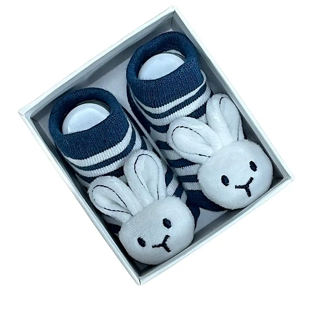 A pair of Baby Bunny Socks by Flowers on Norton St in navy blue, adorned with white stripes and plush white bunny faces, complete with gentle rattles on the toes, are neatly packaged in a white box.