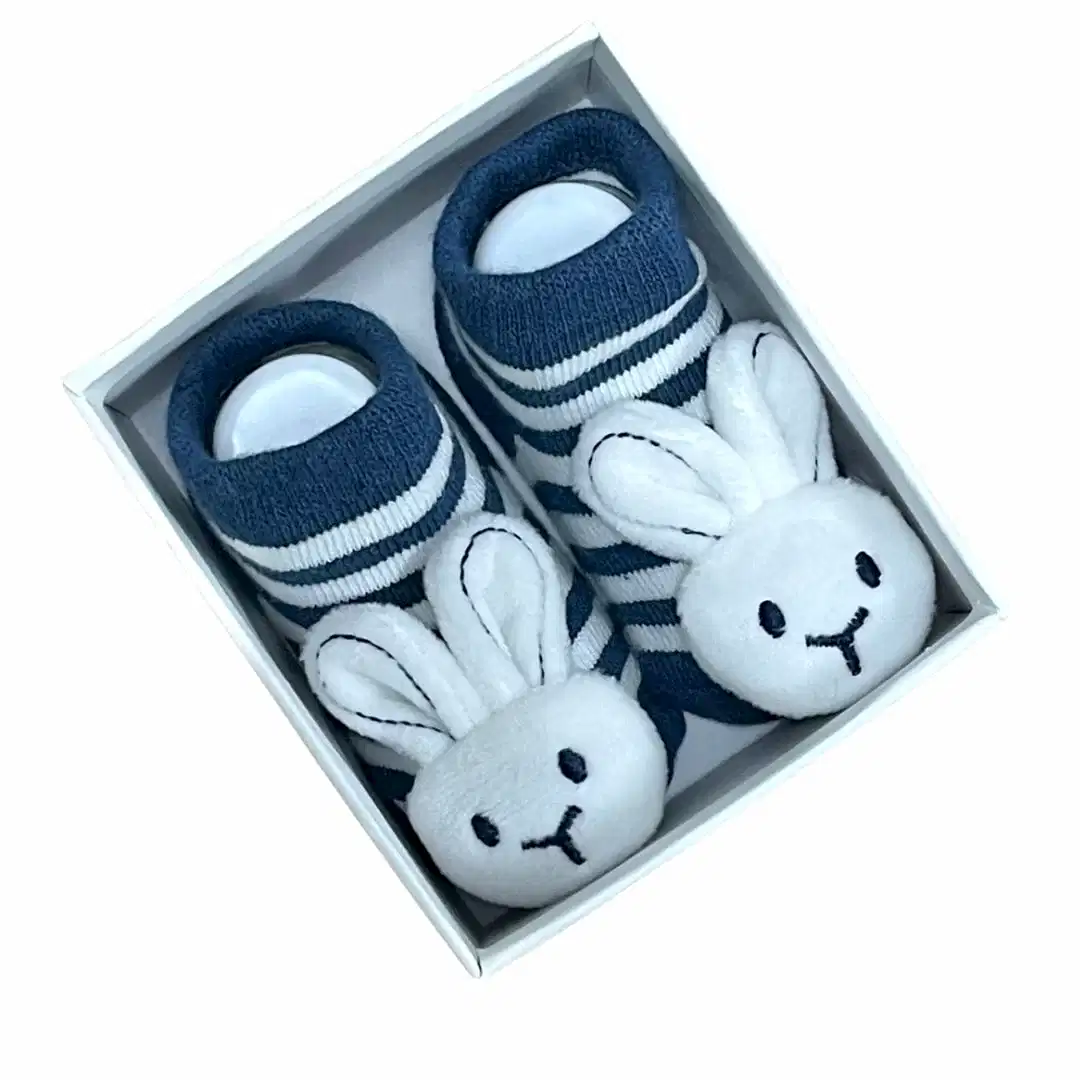 A pair of Baby Bunny Socks by Flowers on Norton St in navy blue, adorned with white stripes and plush white bunny faces, complete with gentle rattles on the toes, are neatly packaged in a white box.