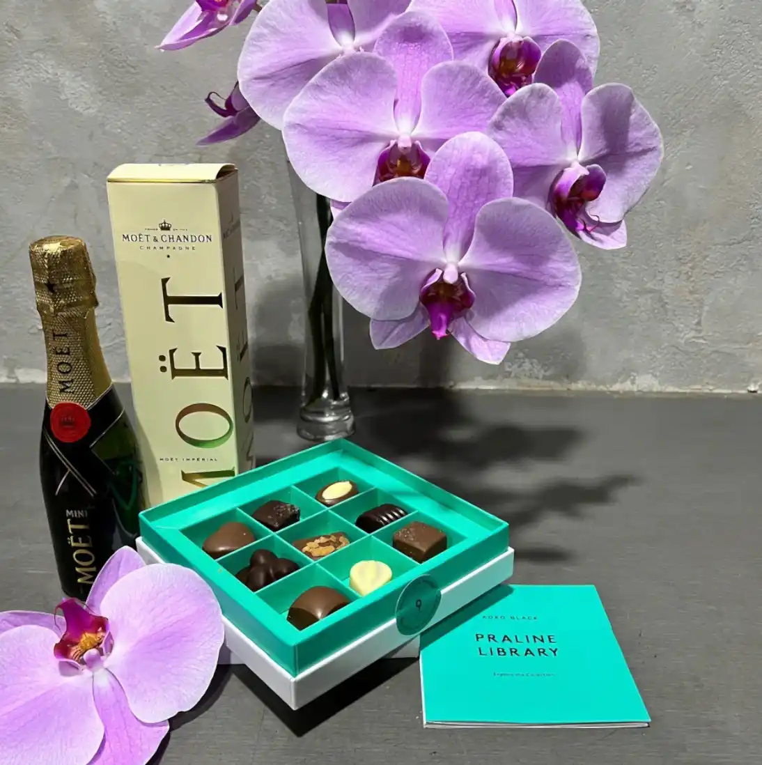 A Little Orchid Delight by Flowers on Norton St graces the table, featuring a box of Koko Black Chocolate Pralines paired with a bottle of Moët champagne. In the background, vibrant purple Phalaenopsis orchids in a vase enhance the elegance, while a teal booklet titled "Praline Library" completes the sophisticated scene.