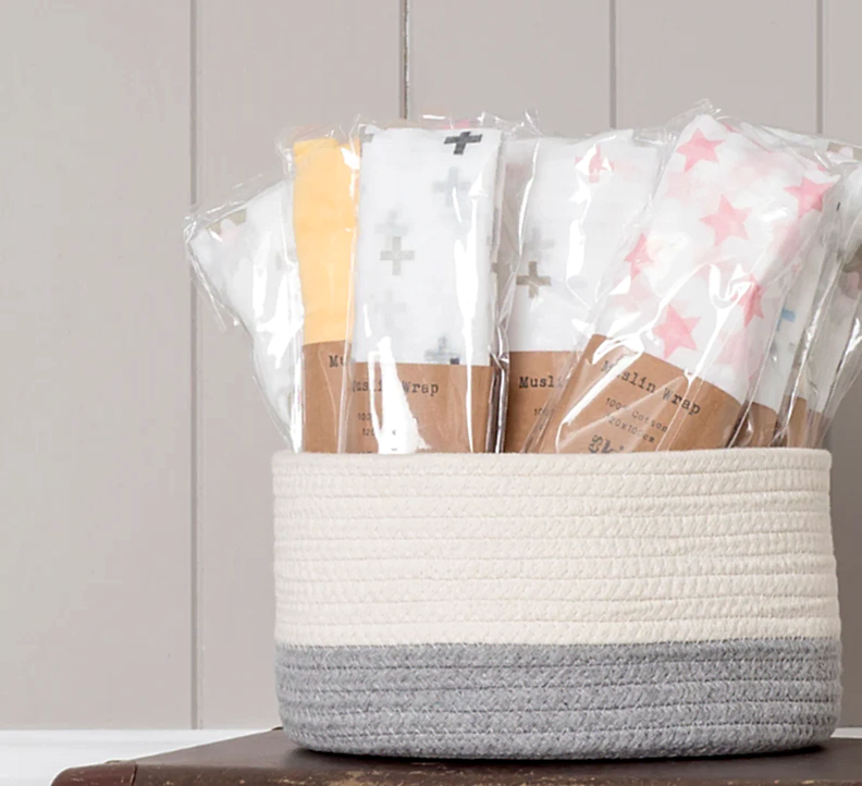 Against a neutral backdrop, a woven basket overflows with FlowersonNortonSt Muslin Wraps in a variety of colors and patterns, such as crosses and stars. These soft wraps are ideal for baby swaddling, offering both comfort and style.