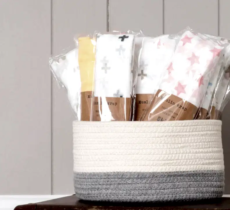 Against a neutral backdrop, a woven basket overflows with FlowersonNortonSt Muslin Wraps in a variety of colors and patterns, such as crosses and stars. These soft wraps are ideal for baby swaddling, offering both comfort and style.