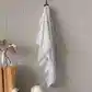 A FlowersonNortonSt Muslin Wrap, featuring a white base with a gray polka dot pattern, hangs on a black hook against a light gray paneled wall. Nearby, a vase holding dried flowers is placed on top of a mustard-colored table draped with cloth. In the foreground, there is part of a chair visible.