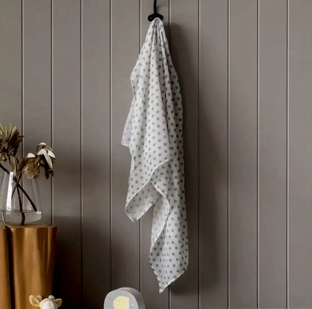 A FlowersonNortonSt Muslin Wrap, featuring a white base with a gray polka dot pattern, hangs on a black hook against a light gray paneled wall. Nearby, a vase holding dried flowers is placed on top of a mustard-colored table draped with cloth. In the foreground, there is part of a chair visible.