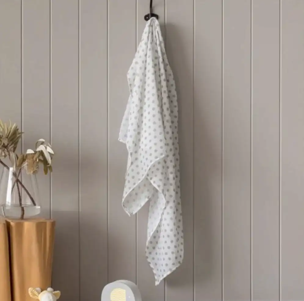 A FlowersonNortonSt Muslin Wrap, featuring a white base with a gray polka dot pattern, hangs on a black hook against a light gray paneled wall. Nearby, a vase holding dried flowers is placed on top of a mustard-colored table draped with cloth. In the foreground, there is part of a chair visible.