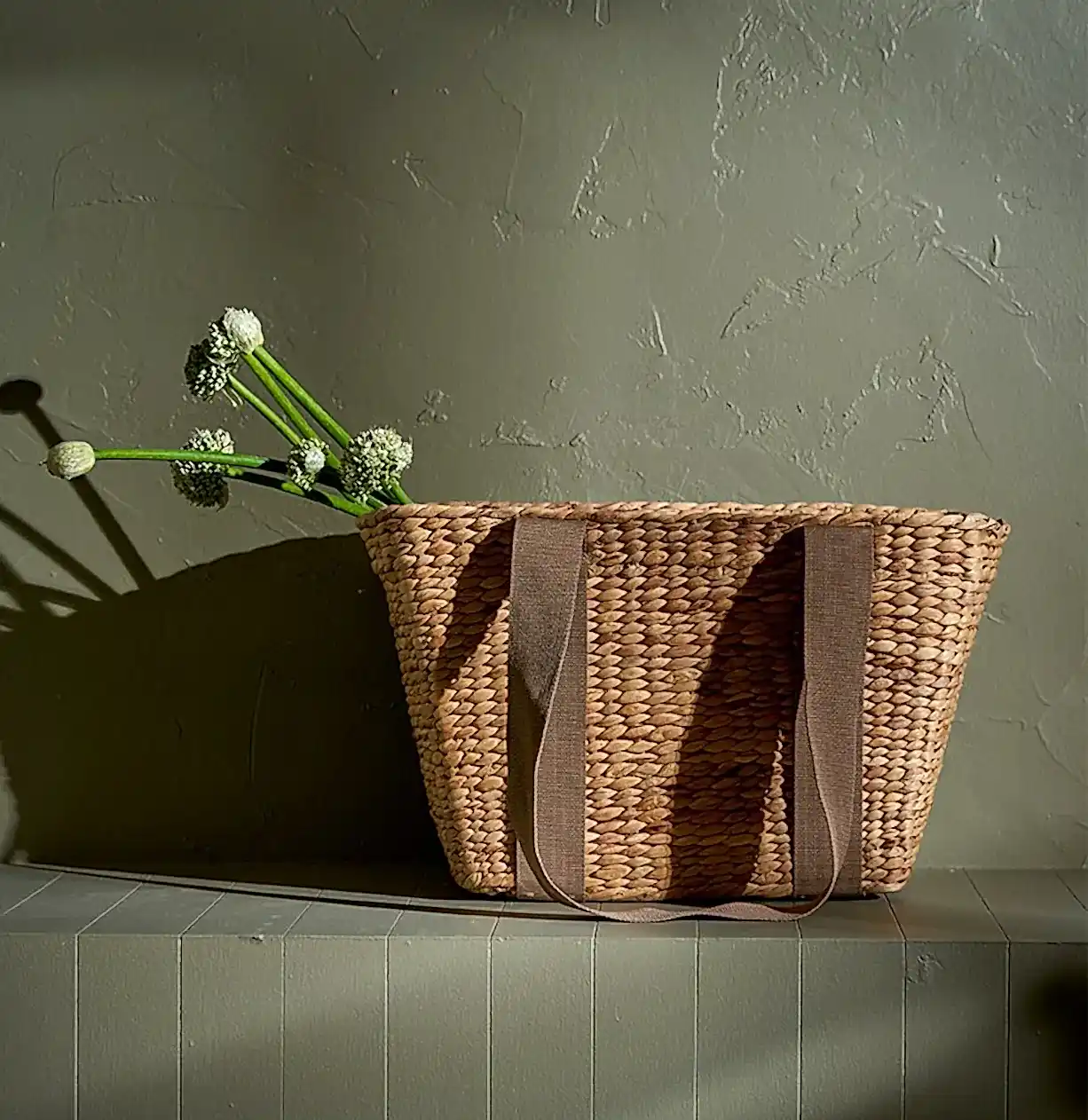 The Journey Tote Bag by Flowers on Norton St, a hand-woven marvel with two sturdy handles, sits on a wooden surface against a textured wall. Long-stemmed white flowers elegantly cascade over one side, casting soft shadows in the warm light.