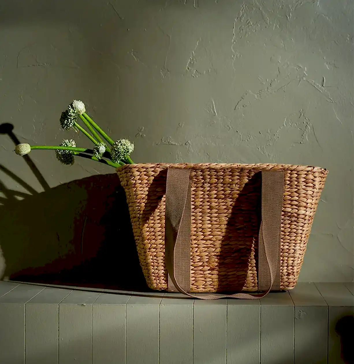 The Journey Tote Bag by Flowers on Norton St, a hand-woven marvel with two sturdy handles, sits on a wooden surface against a textured wall. Long-stemmed white flowers elegantly cascade over one side, casting soft shadows in the warm light.