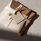 A bundle of brown envelopes, tied with a cream ribbon, labeled "Bath + Body Gift Duo" by Flowers on Norton St, catches the sunlight, casting shadows that highlight the textured paper. The tag notes lavender and lemon myrtle bath products designed for an exfoliating experience.