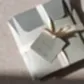 A stack of greenish envelopes tied with cream ribbon rests on a textured surface. A tag reads "Flowers on Norton St - Bath + Body Gift Duo, Unwind." Sunlight casts gentle shadows, inviting you to exfoliate and rejuvenate.