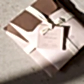 A small stack of pink and brown envelopes tied with a cream ribbon rests on a beige textured surface. The top envelope features a tag labeled "Bath + Body Gift Duo" from Flowers on Norton St, emphasizing its eco-friendly, natural exfoliating qualities.