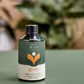 A green bottle labeled "FlowersonNortonSt - We The Wild Plant Care" rests on a lush green surface. This bottle, featuring a plant logo, is filled with organic bioactive plant food and tonic. A softly focused leafy background enhances the natural ambiance, highlighting its commitment to holistic plant care solutions.