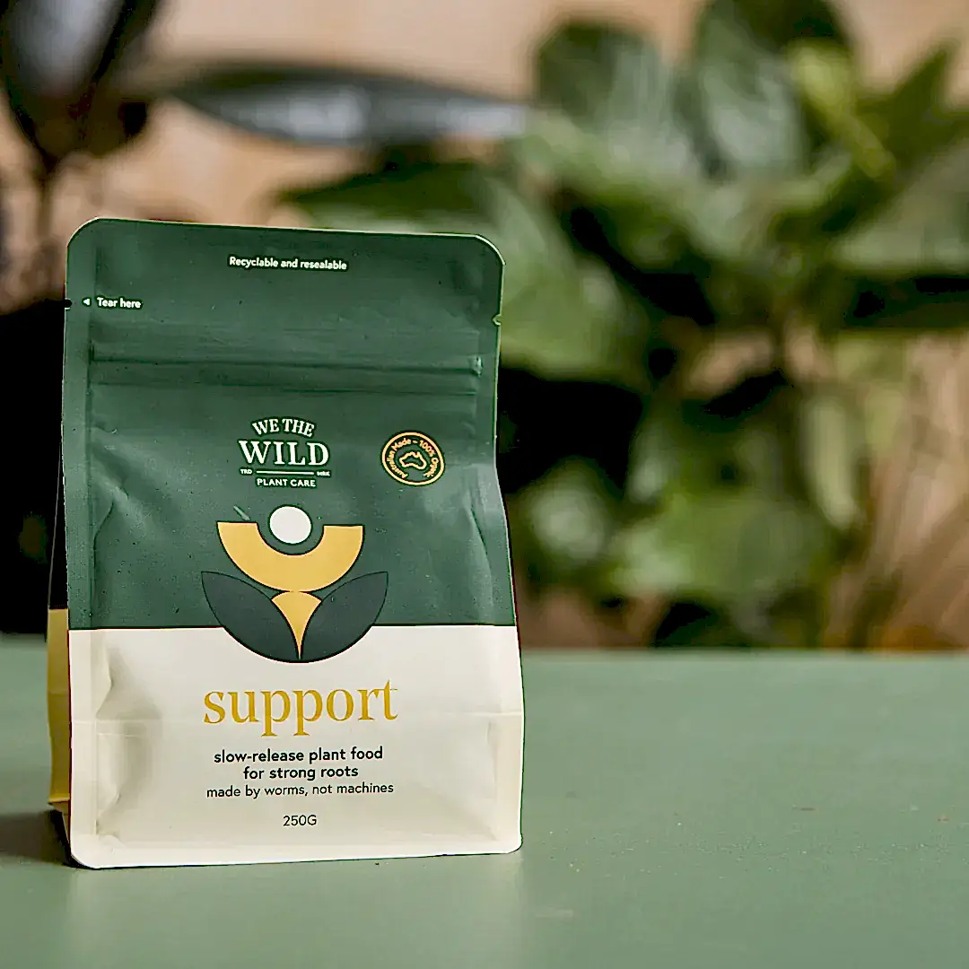 A package of "We The Wild - Plant Care" by FlowersonNortonSt, labeled "Support," contains slow-release plant food made by worms to strengthen roots naturally. The green and beige packaging rests on a green surface, with plant leaves artistically blurred in the background, ideal for naturally tackling pest issues.