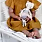 A child in a mustard-yellow dress sits on a white bed, holding a Lily & George | Daisy Dancing Cat doll by Flowers on Norton St. The cat, dressed in a tutu and leotard, features a charming headband with a flower. The child's legs are bare and comfortably stretched out on the bed.