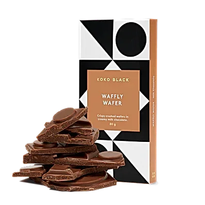 A FlowersonNortonSt Koko Black Chocolate Block wrapped in geometric patterned packaging is displayed beside a stack of broken chocolate pieces. Made with sustainably sourced cocoa, the package notes that it contains crispy crushed wafers in creamy milk chocolate.