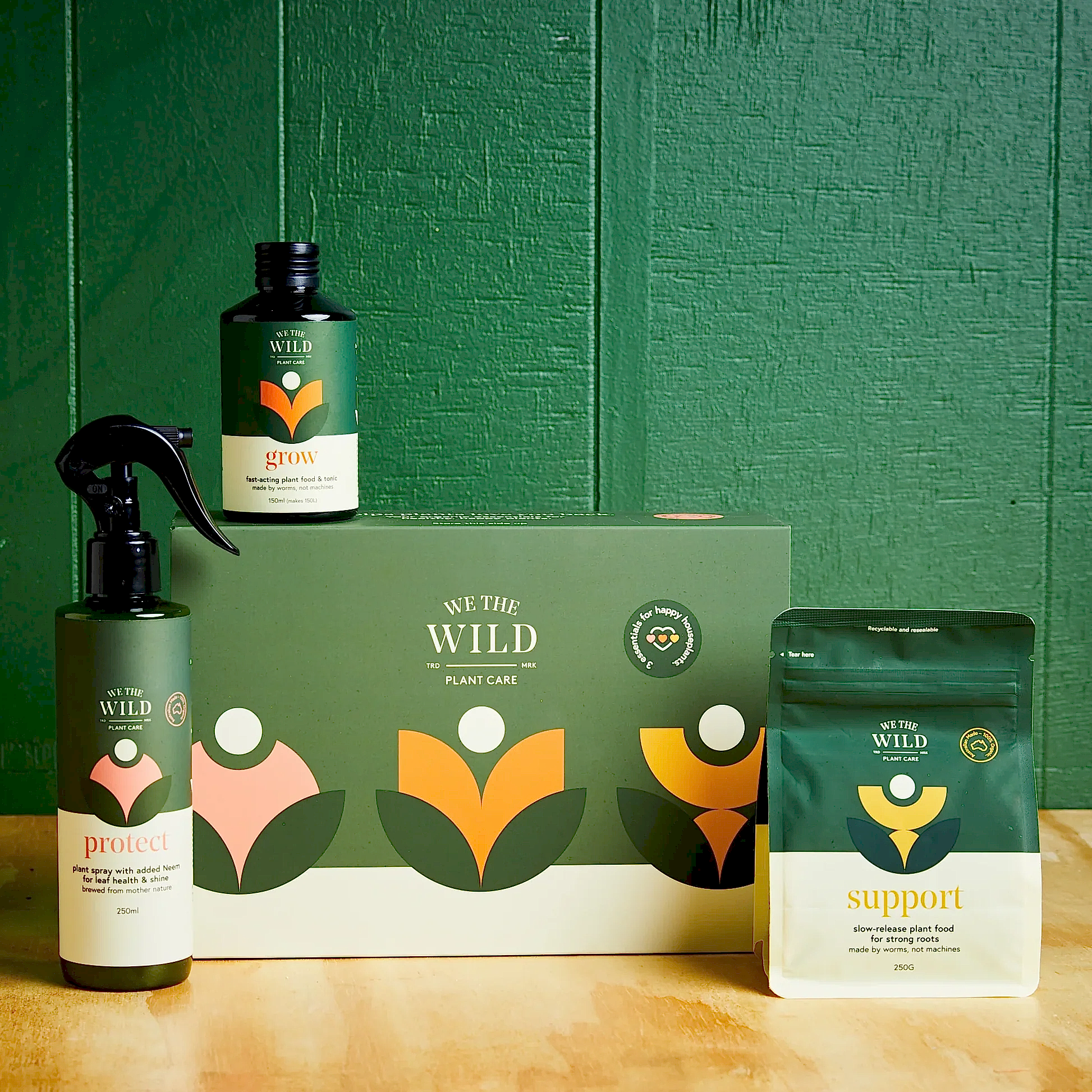Set against a lush green background, a range of FlowersonNortonSt's "We The Wild - Plant Care" products provides effective solutions for pest problems. An artfully arranged collection includes a spray bottle labeled "protect," an organic small bottle labeled "grow," and a pouch titled "support," all surrounding a branded box on a wooden surface.