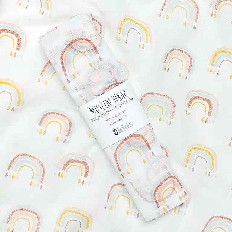 A soft muslin baby wrap adorned with a vibrant rainbow pattern rests on a coordinating background. The wrap is neatly folded and held together with a label that reads "Muslin Wraps, FlowersonNortonSt, 100% Cotton, 120x120cm." The design showcases pastel rainbows in shades of yellow, pink, and blue.