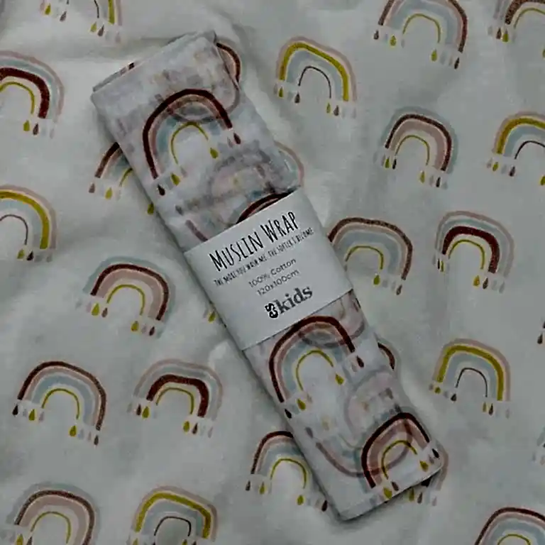 A soft muslin baby wrap adorned with a vibrant rainbow pattern rests on a coordinating background. The wrap is neatly folded and held together with a label that reads "Muslin Wraps, FlowersonNortonSt, 100% Cotton, 120x120cm." The design showcases pastel rainbows in shades of yellow, pink, and blue.